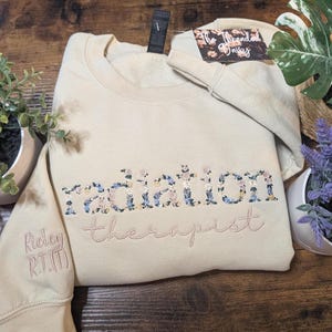 May include: A beige sweatshirt with the words "radiation therapist" embroidered in a floral design. The sleeve has the name "Ridley R.T.(T)" embroidered in a matching color. A tag is attached to the sweatshirt.