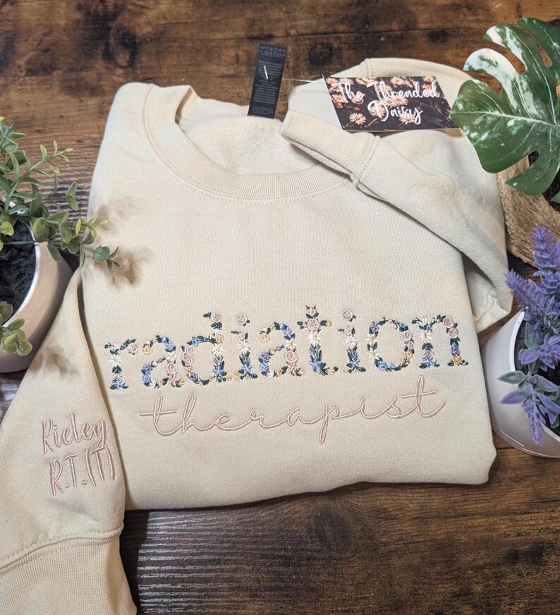 May include: A beige sweatshirt with the words "radiation therapist" embroidered in a floral design. The sleeve has the name "Ridley R.T.(T)" embroidered in a matching color. A tag is attached to the sweatshirt.