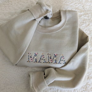 K&ouml;nnte beinhalten: Beiges Sweatshirt mit dem gestickten Wort "MAMA" in floralem Design. Das Sweatshirt ist gefaltet und zeigt den Rundhalsausschnitt und die gerippten &Auml;rmelb&uuml;ndchen. Die Blumenstickerei umfasst rosa, blaue und wei&szlig;e Blumen.