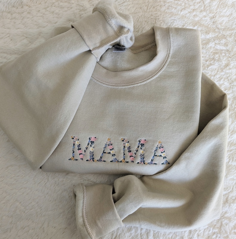 K&ouml;nnte beinhalten: Beiges Sweatshirt mit dem gestickten Wort "MAMA" in floralem Design. Das Sweatshirt ist gefaltet und zeigt den Rundhalsausschnitt und die gerippten &Auml;rmelb&uuml;ndchen. Die Blumenstickerei umfasst rosa, blaue und wei&szlig;e Blumen.