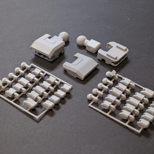 May include: A collection of light gray plastic model kit parts, including several rectangular components and small ball-shaped pieces. Two sprues with numerous small parts are also present, all arranged on a dark surface.