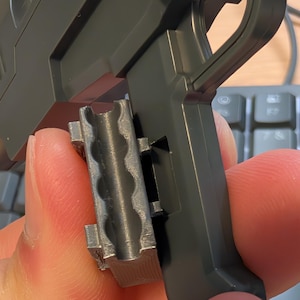 May include: A close-up of a gray plastic toy gun with a handle and a detachable magazine. The magazine is held in place by a finger. The gun is dark gray and has a textured surface.