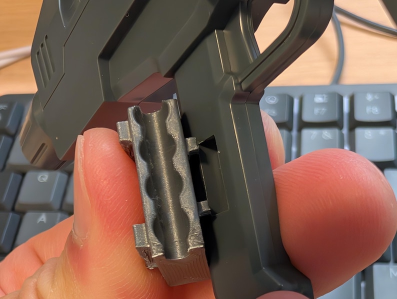 May include: A close-up of a gray plastic toy gun with a handle and a detachable magazine. The magazine is held in place by a finger. The gun is dark gray and has a textured surface.