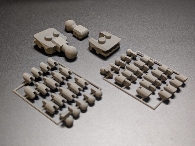 Op de afbeelding: Een verzameling lichtgrijze 3D-geprinte componenten, waaronder verschillende grotere stukken met afgeronde knoppen en kleinere onderdelen gerangschikt in plastic trays. De onderdelen lijken bedoeld te zijn voor een grotere assemblage of model.