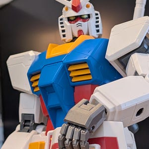 May include: A close-up of a Gundam model, featuring a white head with red and yellow accents, a blue chest, and white arms. The robot has articulated gray hands and is a popular collectible item.