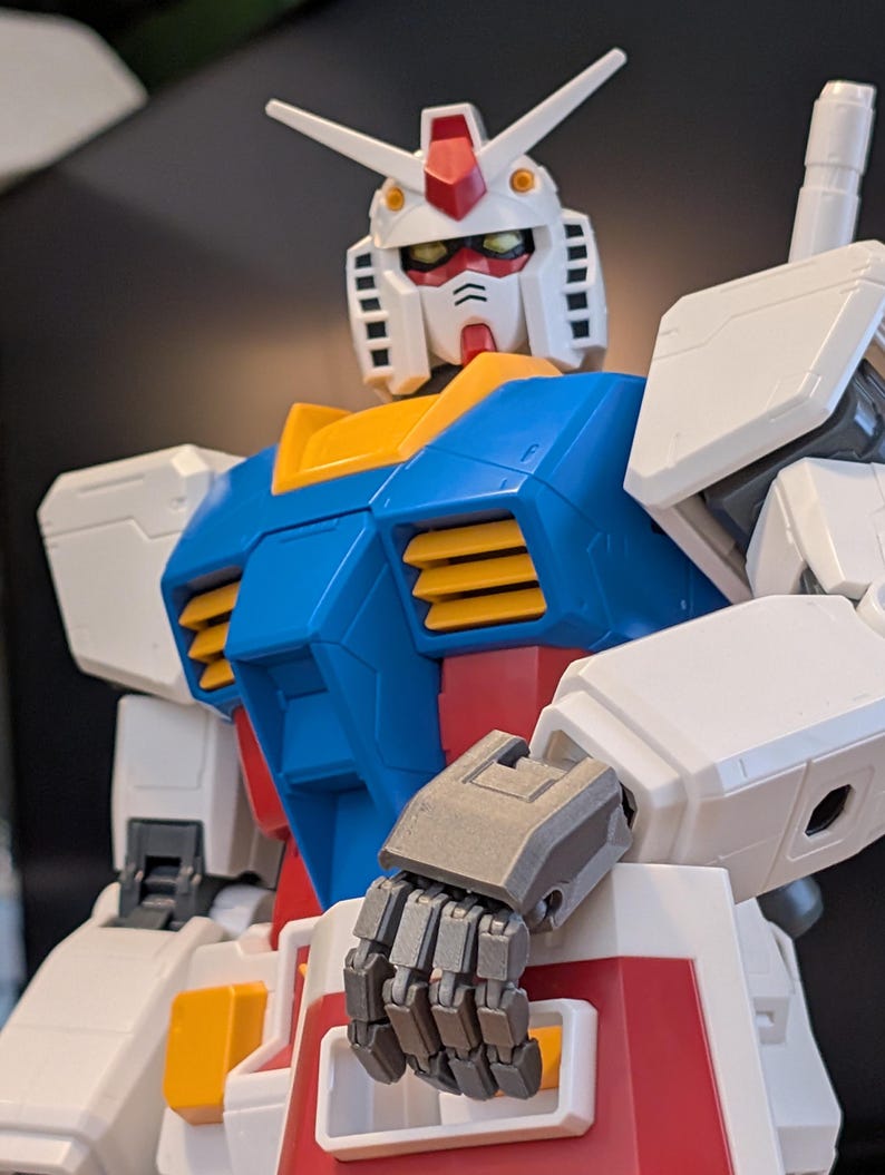 May include: A close-up of a Gundam model, featuring a white head with red and yellow accents, a blue chest, and white arms. The robot has articulated gray hands and is a popular collectible item.