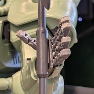 Op de afbeelding: Close-up van een lichtgroene robotfiguur die een grijs wapen vasthoudt. De hand van de robot is gedetailleerd met beweeglijke vingers. De figuur heeft een gladde, matte afwerking. De achtergrond is donker, wat de vorm van de robot benadrukt.