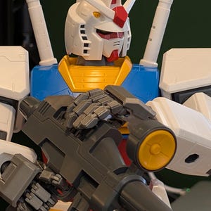 May include: A close-up of a Gundam model, featuring a white head with red and yellow accents. The model has a blue and yellow torso and is holding a gray weapon with a yellow circular detail. The model is a detailed representation of a robot.