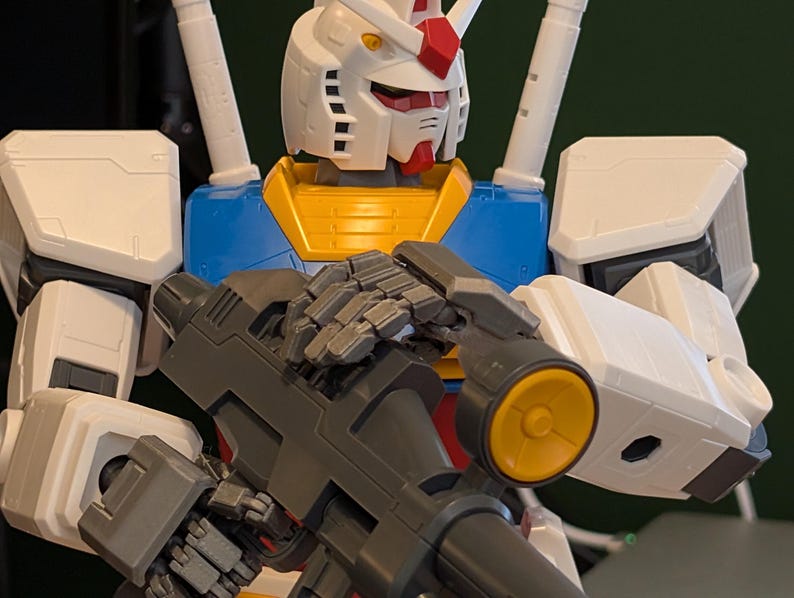 May include: A close-up of a Gundam model, featuring a white head with red and yellow accents. The model has a blue and yellow torso and is holding a gray weapon with a yellow circular detail. The model is a detailed representation of a robot.