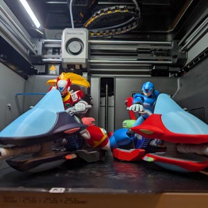 Puede incluir: Dos figuras de acción de personajes de videojuegos, cada una montada en un vehículo futurista, dentro de una impresora 3D. Una figura es roja y amarilla, la otra es azul. Los vehículos tienen detalles en azul claro y rojo.