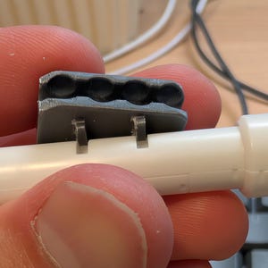 May include: A close-up of a gray plastic component with four circular indentations, attached to a white plastic tube. The component has two small tabs that secure it to the tube. The image is a close-up, showing the details of the plastic parts.