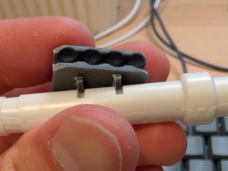 May include: A close-up of a gray plastic component with four circular indentations, attached to a white plastic tube. The component has two small tabs that secure it to the tube. The image is a close-up, showing the details of the plastic parts.