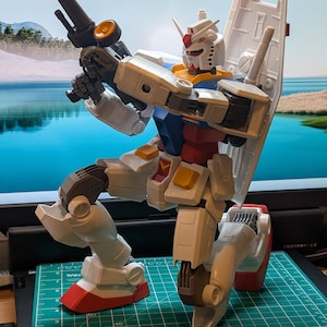 以下が含まれることがあります： 白、青、赤、黄色のガンダムロボットのアクションフィギュアが膝をついたポーズで黒いライフルを構えています。頭部にはV字型のアンテナがあり、背中には大きな白いシールドを背負い、白いグリッド線が入った緑色のカッティングマットの上に置かれています。