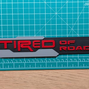 Könnte beinhalten: Ein schwarzes und graues rechteckiges Emblem mit den roten Worten "TIRED OF ROAD". Das Emblem liegt auf einer grünen Schneidematte mit metrischen Maßen.