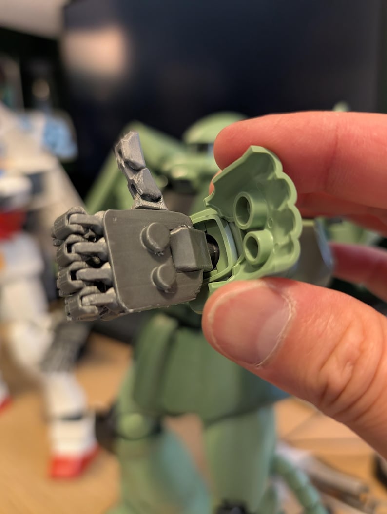 Op de afbeelding: Close-up van een grijze en groene modelbouwkit hand. De hand is gearticuleerd en lijkt deel uit te maken van een groter robotmodel. De hand wordt tegen een groen plastic stuk gehouden, mogelijk de arm.