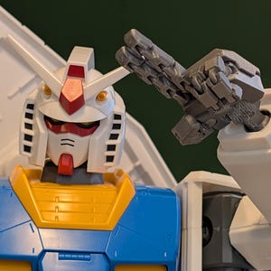 May include: A close-up of a Gundam model, featuring a white head with red and yellow accents, and a blue and yellow torso. The robot's articulated arm is raised, showcasing its detailed mechanical design. The model is a collectible item.