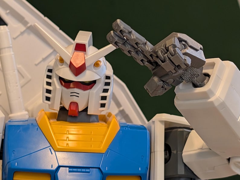May include: A close-up of a Gundam model, featuring a white head with red and yellow accents, and a blue and yellow torso. The robot's articulated arm is raised, showcasing its detailed mechanical design. The model is a collectible item.