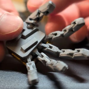 May include: A gray articulated robotic hand with multiple jointed fingers. The hand is held by a person. The fingers are made of small, rectangular segments. The base of the hand is a solid gray color with yellow accents.