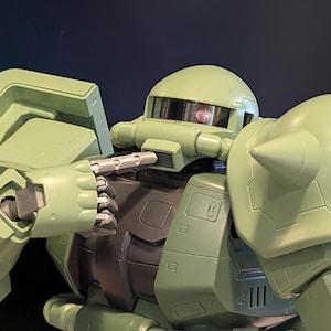 Op de afbeelding: Een close-up van een groene robotactiefiguur. De figuur heeft een afgeronde helm met een vizier, een cilindrische nek en een grote schouder met een conische punt. De hand van de robot houdt een zilveren voorwerp vast.
