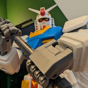 May include: A close-up of a Gundam model, featuring a white, blue, yellow, and red color scheme. The robot holds a gray weapon. The model has a detailed design with visible mechanical parts and is posed in a dynamic stance.