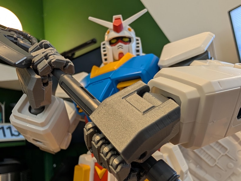 May include: A close-up of a Gundam model, featuring a white, blue, yellow, and red color scheme. The robot holds a gray weapon. The model has a detailed design with visible mechanical parts and is posed in a dynamic stance.