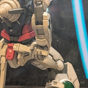 May include: A detailed robot action figure in white, red, navy blue, and grey. The figure is posed dynamically, with intricate mechanical details on its limbs and torso. A blue light effect is in the background. The figure is a collectible item.