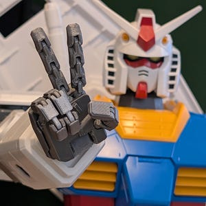 May include: A close-up of a Gundam model kit, a Japanese mecha robot, making a peace sign with its articulated hand. The robot has a white head with red and yellow accents, blue and yellow torso, and gray articulated hand.