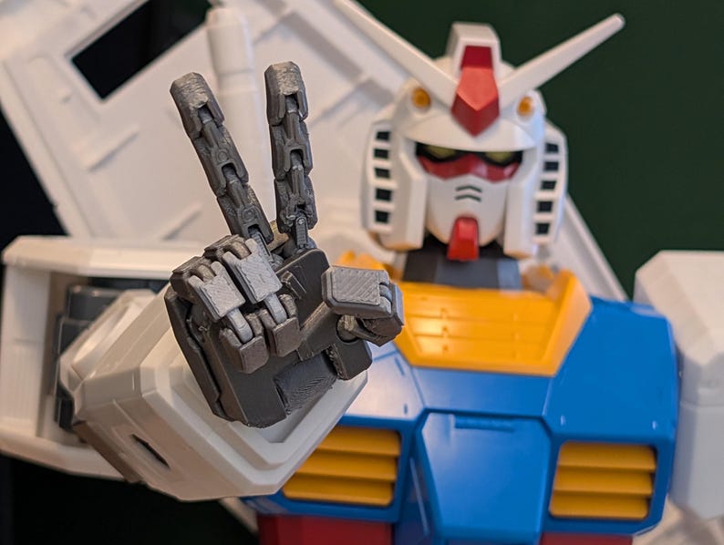 May include: A close-up of a Gundam model kit, a Japanese mecha robot, making a peace sign with its articulated hand. The robot has a white head with red and yellow accents, blue and yellow torso, and gray articulated hand.