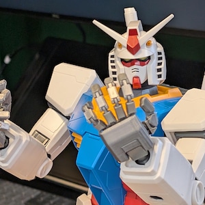 May include: A detailed, close-up view of a Gundam model. The robot features a white head with red and yellow accents, blue and yellow torso, and white arms. The hands are open, displaying intricate mechanical details. The model is a collectible item.