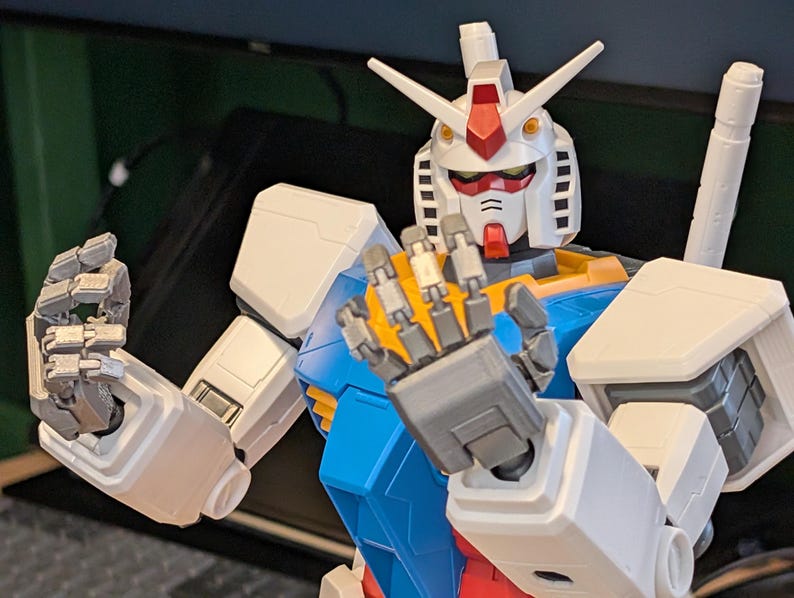 May include: A detailed, close-up view of a Gundam model. The robot features a white head with red and yellow accents, blue and yellow torso, and white arms. The hands are open, displaying intricate mechanical details. The model is a collectible item.