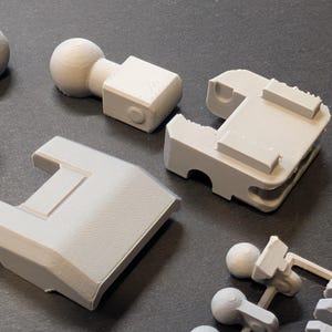 May include: Close-up of several light gray 3D-printed plastic components. The parts include ball joints, rectangular blocks, and other geometric shapes. The objects are arranged on a dark surface, showcasing their design and texture.