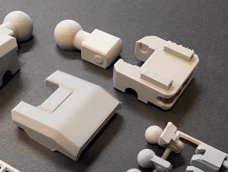 May include: Close-up of several light gray 3D-printed plastic components. The parts include ball joints, rectangular blocks, and other geometric shapes. The objects are arranged on a dark surface, showcasing their design and texture.
