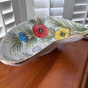 May include: A white ceramic bowl with a gold rim and a floral design. The bowl has a crescent shape and features red, yellow, and blue flowers with green leaves.