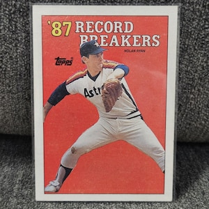Pode incluir: Cartão de beisebol com Nolan Ryan em uniforme dos Astros. O cartão tem um fundo vermelho com o texto "'87 Record Breakers" no topo. O logotipo da Topps é visível. O jogador está em pose de arremesso.