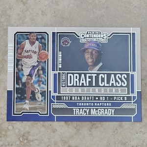 Pode incluir: Um cartão de basquete Panini Contenders com Tracy McGrady do Toronto Raptors. O cartão mostra McGrady com o uniforme dos Raptors e um terno. O texto inclui "Draft Class" e "1997 NBA Draft".