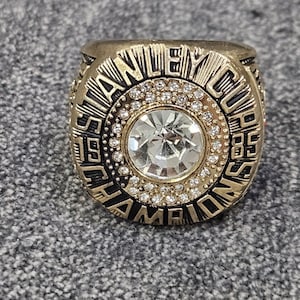 以下が含まれることがあります： 「STANLEY CUP CHAMPIONS 85」の文字が刻まれたゴールドトーンのリング。リングの中央には、大きな透明なファセットストーンがあり、その周りを小さな透明なストーンが囲んでいます。