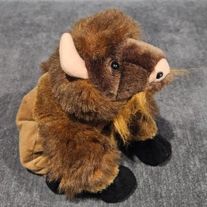 May include: A plush bison toy featuring thick, dark brown fur on the head and shoulders, lighter brown fur on the body, black hooves, and light tan horns. The stuffed animal has a small beard of longer, golden-brown fibers under the chin.