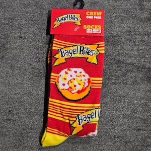 Puede incluir: Un par de calcetines rojos de corte medio con rayas horizontales amarillas y un logotipo repetido de Bagel Bites con un gráfico de un bagel de pizza. El encabezado del empaque es rojo con el logo de Bagel Bites y el texto Crew One Pair Socks.