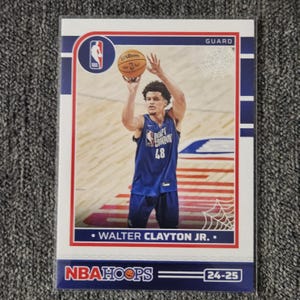 Puede incluir: Una tarjeta de baloncesto con Walter Clayton Jr. con una camiseta azul con el número 48, lanzando un balón de baloncesto. La tarjeta tiene un borde blanco con detalles en rojo y azul, el logo de NBA Hoops y el texto "GUARD".