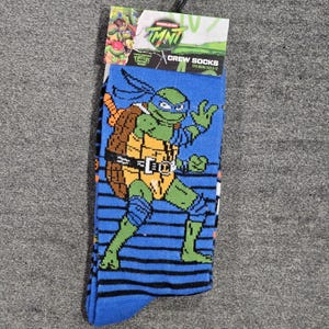 Könnte beinhalten: Blaue Crew-Socken mit einem Teenage Mutant Ninja Turtle-Charakter. Die Socken haben einen blauen Hintergrund mit schwarzen Streifen und eine Grafik des Charakters in Grün, Braun und Gelb. Die Verpackung trägt die Aufschrift "TMNT Crew Socks".