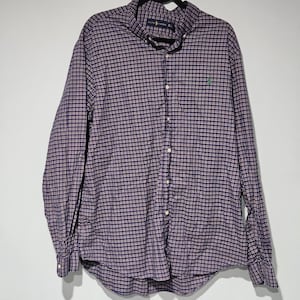 May include: A long-sleeved button-down shirt with a plaid pattern in shades of purple, white, and red. The shirt has a button-down collar and a small green logo on the chest. The shirt is hanging on a black hanger.