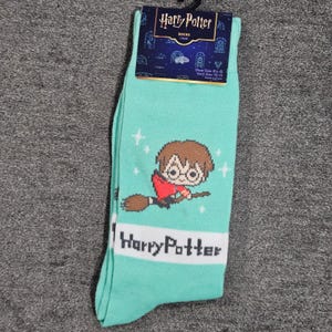 May include: A pair of mint green Harry Potter socks. The socks feature a cartoon image of Harry Potter on a broomstick, with the words "Harry Potter" in white block letters. The socks have a tag at the top with the Harry Potter logo.