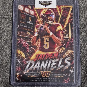 May include: A sports trading card featuring Jayden Daniels of the Washington Commanders in a burgundy and gold uniform, holding a football. The card displays the text Jayden Daniels, Washington Commanders, Quarterback 1/5, and Drumbum Designs 2026.
