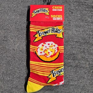 May include: A pair of red crew socks featuring yellow horizontal stripes and a repeating Bagel Bites logo with a graphic of a pizza bagel. The packaging header is red with white text that reads Bagel Bites Crew One Pair Socks Fits Men's Shoe Size 6-12.