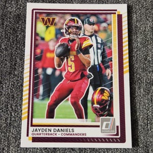 May include: A Donruss football trading card featuring Jayden Daniels, quarterback for the Commanders, in a burgundy and gold uniform. The card has a white border with burgundy and gold accents. The Donruss logo is visible.