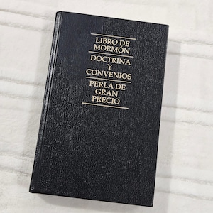 May include: A black hardcover book with gold lettering. The text on the cover reads "LIBRO DE MORMON, DOCTRINA Y CONVENIOS, PERLA DE GRAN PRECIO". The book is closed and appears to be in good condition.