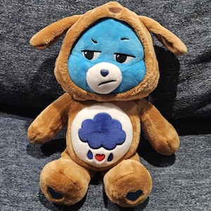 May include: A plush toy featuring a blue bear face with a grumpy expression, wearing a brown dog-themed hood with floppy ears. The bear has a white belly patch displaying a dark blue rain cloud with a small red heart and two blue raindrops.