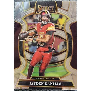 May include: A 2025 Select football trading card featuring Jayden Daniels in a burgundy and gold Commanders uniform. The player is shown in a throwing stance holding a football. The card has a metallic finish with the text Select 2025 and Commanders.