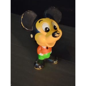 May include: Vintage Mickey Mouse figurine. The figure has a yellow face, black ears, and a red shirt with green shorts. The figure is standing on a black surface.