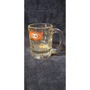 May include: A clear glass mug with a thick handle and a dimpled base. The exterior features an orange graphic of the United States map with the white and brown A&W logo centered within the map outline, printed clearly on the side of the glass.